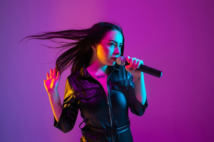 Foto retrato de cantante de raza caucásica aislado sobre fondo de estudio púrpura en luz de neón. modelo de mujer hermosa en ropa negra con micrófono. concepto de emociones humanas, expresión facial, publicidad, música, arte.