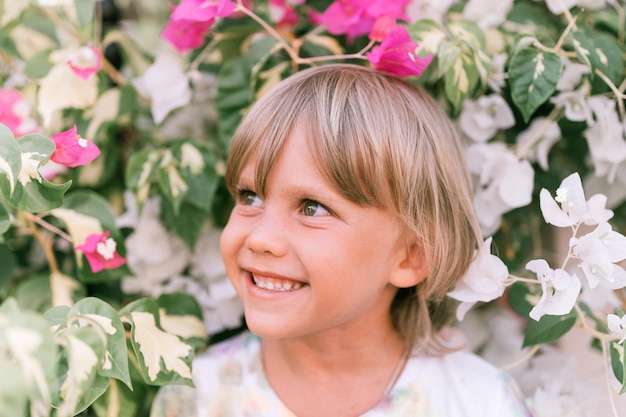 Retrato de la cara de un niño rubio de cinco años de edad, feliz y sonriente, con ojos verdes en plantas de flores rosas y blancas en la naturaleza, los niños se divierten las vacaciones de verano, luminoso y aireado