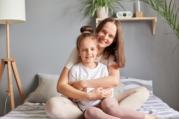 Retrato casero de una mujer sonriente y una niña pequeña madre e hija sentadas en la cama abrazándose en el dormitorio mirando a la cámara expresando felicidad felicitando el día de la madre