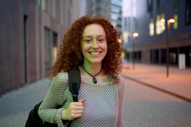 Retrato caucásica joven estudiante pelirroja sonriendo mirando feliz a la cámara calle noche al aire libre