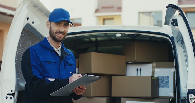 Retrato del caucásico apuesto trabajador de la compañía naviera de pie en la camioneta con cajas y escribiendo el documento, luego sonriendo a la cámara. Al aire libre.