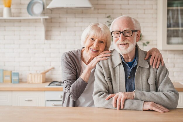 Foto retrato de caucásico sonriente anciano anciano pareja familia cónyuges abuelos mirando a la cámara abrazando abrazando con amor y cuidado en la cocina de casa