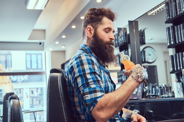 Retrato de cerca de un apuesto y elegante hombre barbudo con un tatuaje en el brazo vestido con una camisa de franela bebe jugo en una barbería.