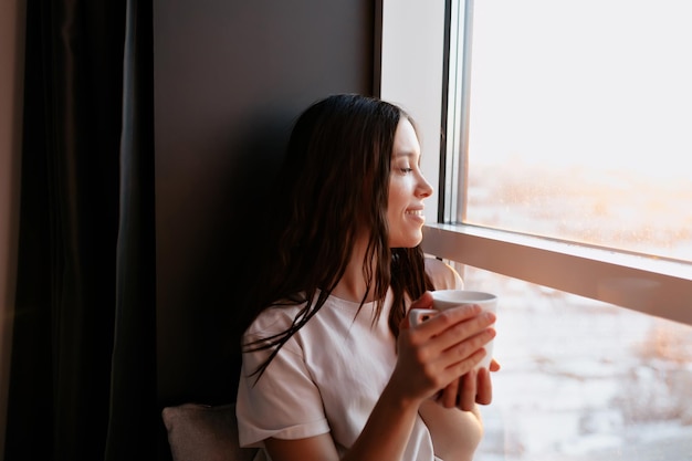 Retrato de cerca de una chica feliz y encantadora con cabello oscuro sosteniendo café y mirando la ventana con una sonrisa feliz Nuevo día en casa con sol Solo en casa