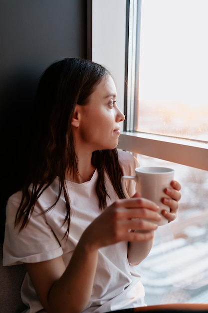 El retrato de cerca de una chica de pelo oscuro con camiseta blanca sostiene una taza con café y mira por la ventana con una sonrisa encantadora por la mañana