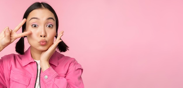 El retrato de cerca de una elegante chica asiática muestra un signo de paz y un beso posando sobre un fondo rosa