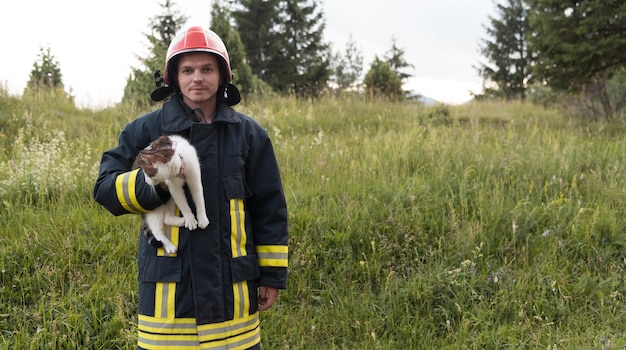 Retrato de cerca de un heroico bombero con traje protector y casco rojo que sostiene a un gato salvado en sus brazos. Bombero en operación de extinción de incendios. foto de alta calidad