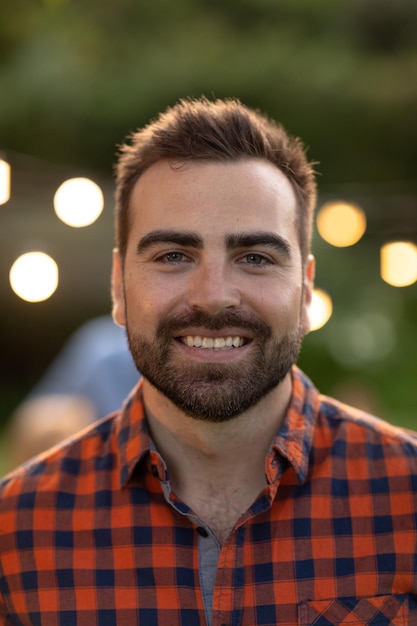 Retrato de cerca de un hombre caucásico con cabello oscuro corto y barba, usando una camisa a cuadros en el jardín sonriendo a la cámara, luces bokeh en el fondo