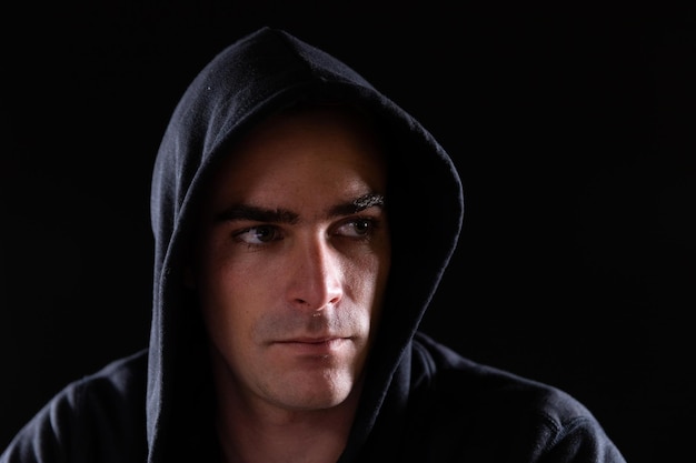 Retrato de cerca de un hombre caucásico preocupado que llevaba una sudadera con capucha negra trabajando hasta tarde en la oficina mirando de reojo sobre un fondo negro.