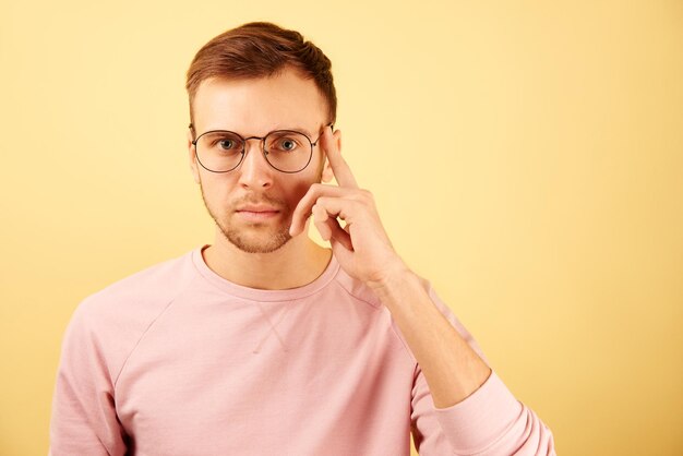 El retrato de cerca de un hombre inteligente que usa anteojos con la cara enfocada muestra los dedos en su sien recomienda pensar aislado en un fondo amarillo con ropa informal rosa