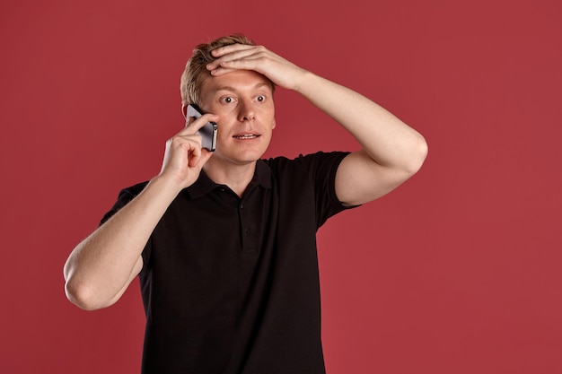 Retrato de cerca de un joven atlético pelirrojo con un elegante pantalones negro hablando por un smartphone mientras posa en un estudio rosa. Expresiones faciales humanas. Concepto de emociones sinceras. Copiar