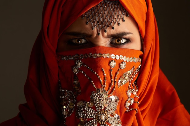 Retrato de cerca de una joven enojada con encantadores ojos ahumados y joyas en la frente, usando el hiyab de terracota decorado con lentejuelas. Ella está mirando a la cámara sobre un fondo oscuro. H