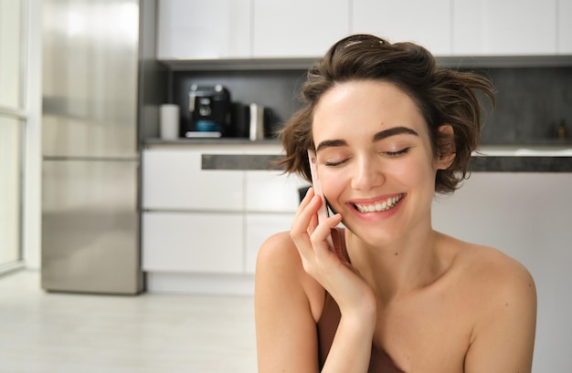 Retrato de cerca de una mujer feliz y sonriente hablando por teléfono móvil desde su cocina con una chica charlatana