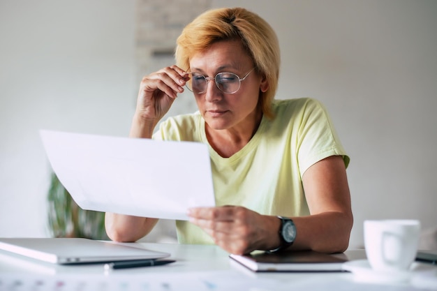 Retrato de cerca de una mujer inteligente de mediana edad con gafas de protección con papeles mientras está sentada en el lugar de trabajo frente a la computadora portátil en la oficina de casa