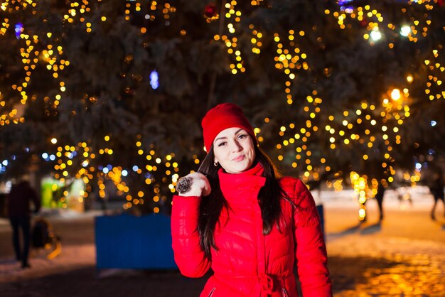 Retrato cercano de una atractiva modelo joven con un abrigo rojo brillante y mitones de lana parados frente al árbol de Navidad de la ciudad con luces encendidas. Concepto de vacaciones de invierno