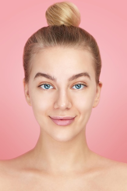 Retrato cercano de una joven y bella mujer rusa de ojos azules desnuda sin maquillaje sonriendo suavemente con un moño de cabello rubio en la cabeza aislado en rosa, belleza natural