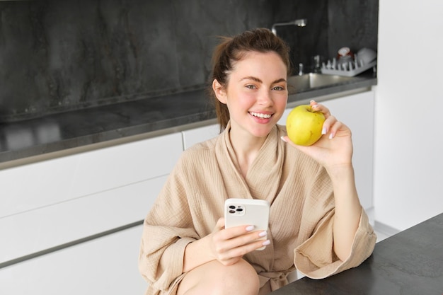 Un retrato cercano de una mujer morena sonriente en bata de baño se sienta en la cocina de casa usa teléfono móvil