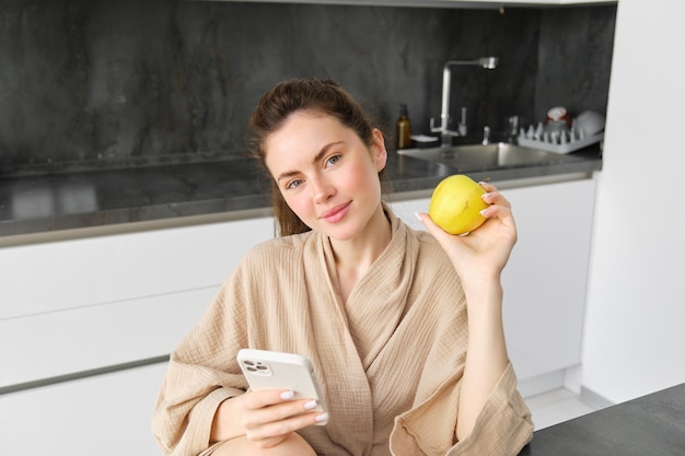 Un retrato cercano de una mujer morena sonriente en bata de baño se sienta en la cocina de casa usa teléfono móvil