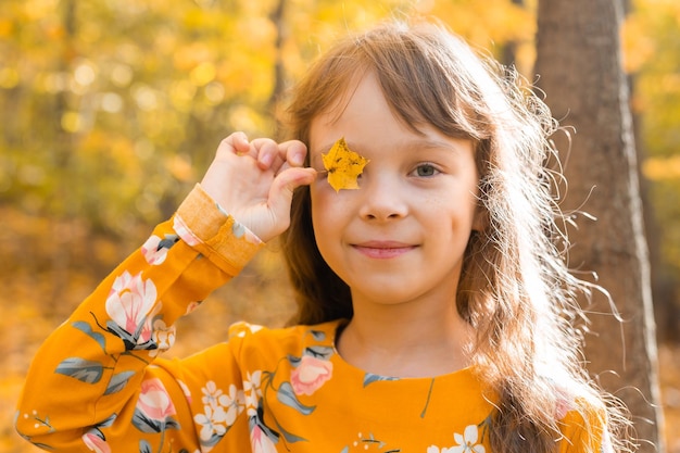Retrato cercano de una niña que cubre su ojo con una hoja de otoño de arce Temporada de otoño y concepto de niños