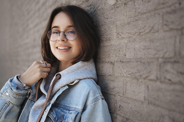 Retrato cerrado de una alegre y sonriente chica de pelo corto con gafas y chaqueta de mezclilla apoyada en la pared del edificio mirando a la cámara con expresión feliz y despreocupada disfrutando de caminar afuera