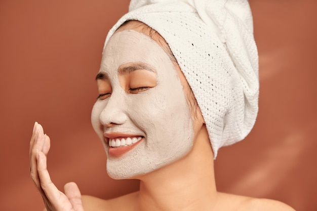 Retrato cerrado de una hermosa chica asiática en un spa con una toalla en la cabeza aplicando una máscara facial de arcilla