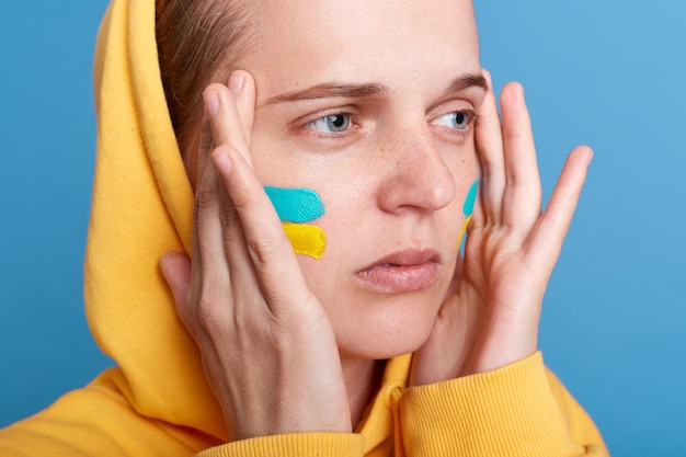 Retrato cerrado de una mujer triste con capucha con la bandera ucraniana en las mejillas posando aislada sobre fondo azul, una mujer patriótica que expresa tristeza se preocupa por Ucrania y mantiene las manos en los templos