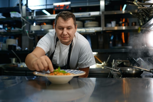 Retrato de un chef satisfecho colocando hojas de albahaca en un plato con pasta recién cocinada, enfoque selectivo. Comida tradicional, concepto de alimentación saludable.