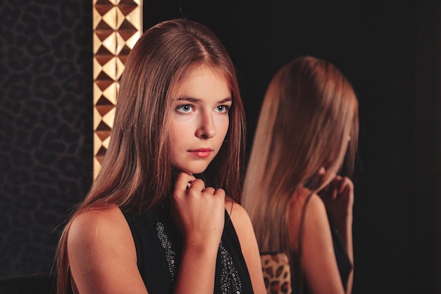 Retrato de una chica adolescente muy linda con un vestido elegante en el espejo en el interior oscuro con estilo de la sala de estar. Niños emociones y poses. Concepto de estilo, moda y belleza. Copie el espacio para el sitio