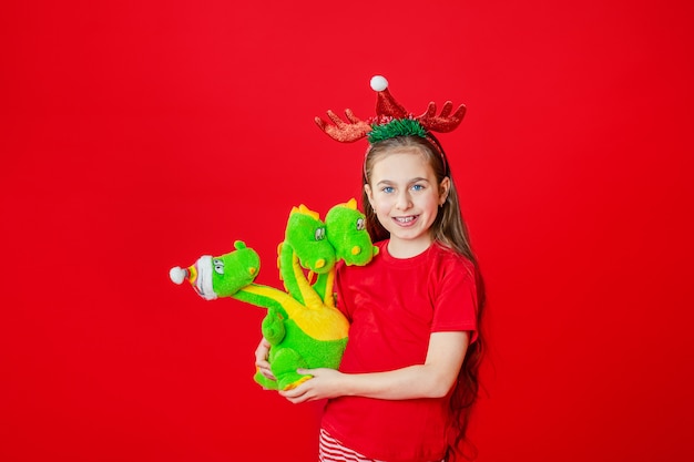 Retrato de una chica alegre divertida con un vendaje de cuernos en la cabeza abrazando a un dragón de peluche en pijama de Navidad aislado