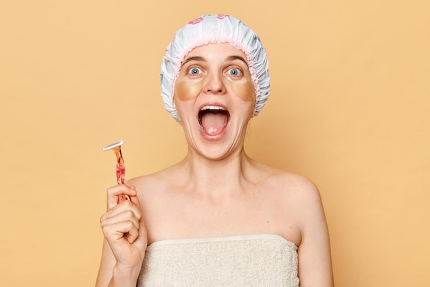 Retrato de una chica alegre y emocionada con gorro de ducha sosteniendo en la mano una cuchilla de afeitar aislada sobre fondo beige gritando de pie con la boca abierta