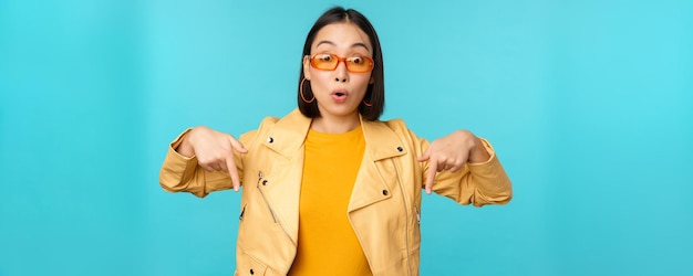 El retrato de una chica asiática con estilo usa gafas de sol, sonríe y señala con el dedo hacia abajo, muestra anuncios sobre fondo azul.