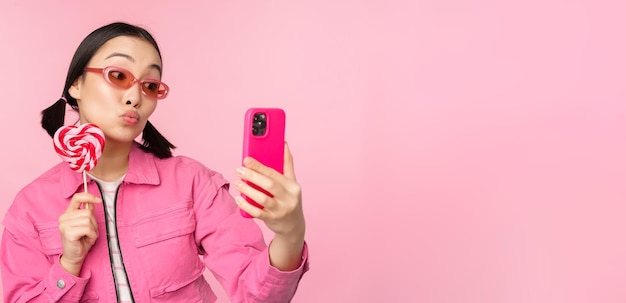 Retrato de una chica asiática feliz y elegante que se toma una selfie con dulces de lolipop y sonríe tomando una foto con una aplicación móvil de pie sobre un fondo rosa