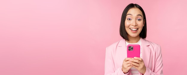 Retrato de una chica asiática en traje con teléfono inteligente sonriendo y luciendo feliz de pie sobre fondo de estudio rosa