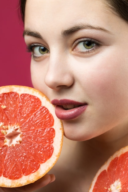 Retrato de una chica atractiva sosteniendo una rodaja de pomelo sobre un fondo rojo.