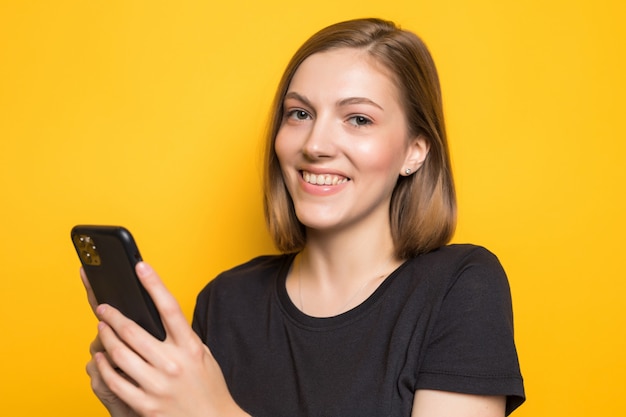 Retrato de chica atractiva sosteniendo un teléfono inteligente en las manos, revisando el correo electrónico, usando internet 5G, escribiendo sms, de pie sobre una pared amarilla