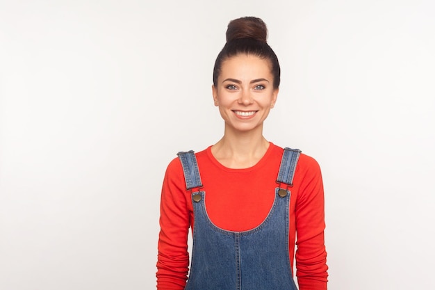 Retrato de una chica bonita, feliz y elegante, con un moño en overoles de mezclilla, mirando a la cámara con una sonrisa atractiva, expresando alegría y optimismo. Foto de estudio aislado sobre fondo blanco.