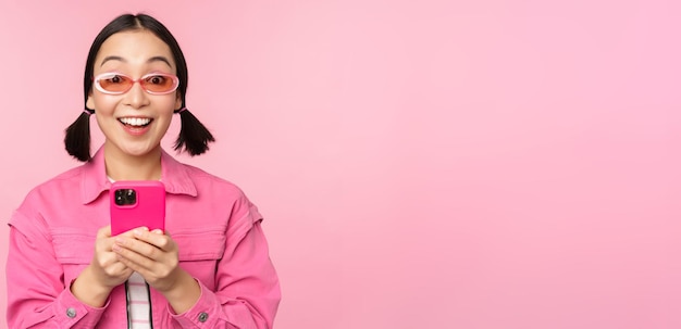 Retrato de una chica coreana con elegantes gafas de sol sosteniendo un teléfono móvil usando una aplicación de teléfono inteligente de pie sobre fondo rosa Copiar espacio