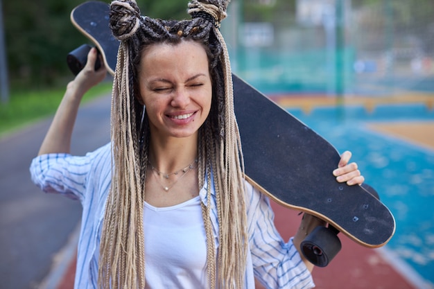 Retrato de una chica divertida con los ojos cerrados y pequeñas coletas en la cabeza que sostiene un longboard b ...