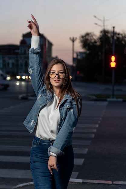 Retrato de una chica enérgica con ropa de mezclilla en el fondo de la ciudad por la noche Mujer joven elegante con gafas en la calle de la ciudad