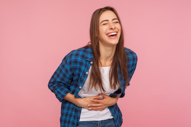 Retrato de una chica extremadamente feliz con pantalones a cuadros sosteniendo su estómago y riéndose a carcajadas, riéndose de una anécdota divertida, emoción sincera. tiro de estudio interior aislado sobre fondo de color rosa