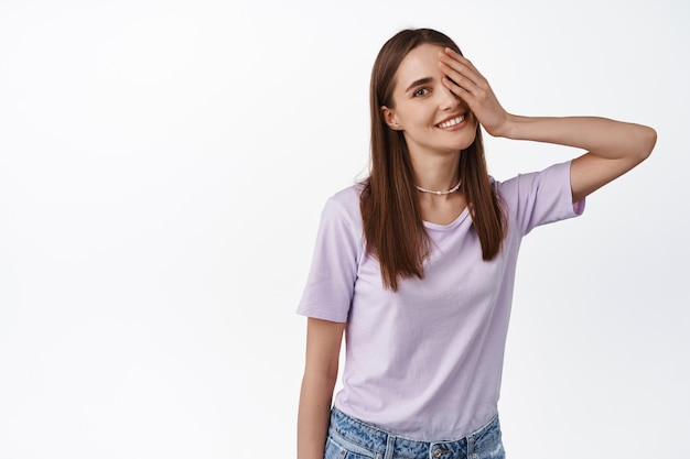 Foto retrato de una chica feliz y hermosa que cubre un lado de la cara y mira la cámara con una linda sonrisa, parada despreocupada contra un fondo blanco con una camiseta morada.