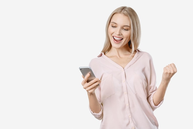 Retrato de una chica feliz sosteniendo un teléfono móvil y celebrando la victoria aislada sobre fondo blanco