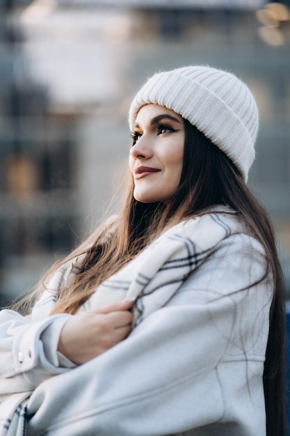 Retrato de una chica guapa soñando sonriendo en el fondo de la ciudad Vista lateral de una chica hermosa vestida con sombrero blanco y bufanda a cuadros