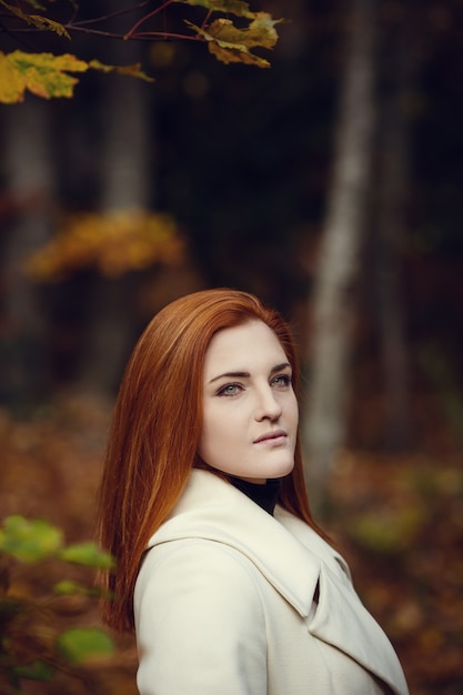 Retrato de una chica hermosa, soñadora y triste con el pelo rojo en otoño de bata blanca