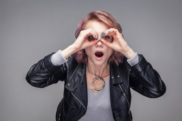 Retrato de una chica hermosa sorprendida con peinado corto y maquillaje en una chaqueta de cuero negro de estilo casual de pie y mirando con gesto binocular. tiro de estudio interior, aislado sobre fondo gris.