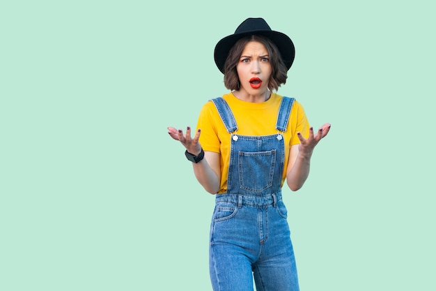 Retrato de chica hipster bastante joven sorprendida en overoles de mezclilla azul, camisa amarilla y sombrero negro de pie con los brazos levantados y mirando a cámara. tiro del estudio de interior aislado en fondo verde claro.