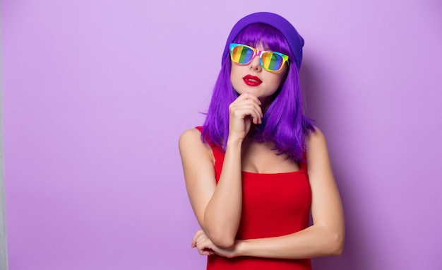 Retrato de una chica hipster de estilo joven con cabello morado y anteojos de arco iris sobre fondo rosa