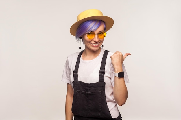 Retrato de una chica hipster glamurosa moderna con el pelo corto violeta con gafas de sol y sombrero sonriendo y señalando hacia un lado, copiando espacio para publicidad. aislado sobre fondo blanco, tiro de estudio interior