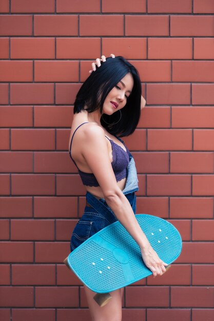 Retrato de chica hipster con monopatín sobre fondo de pared de ladrillo. Hermosa mujer asiática posa para tomar una foto.