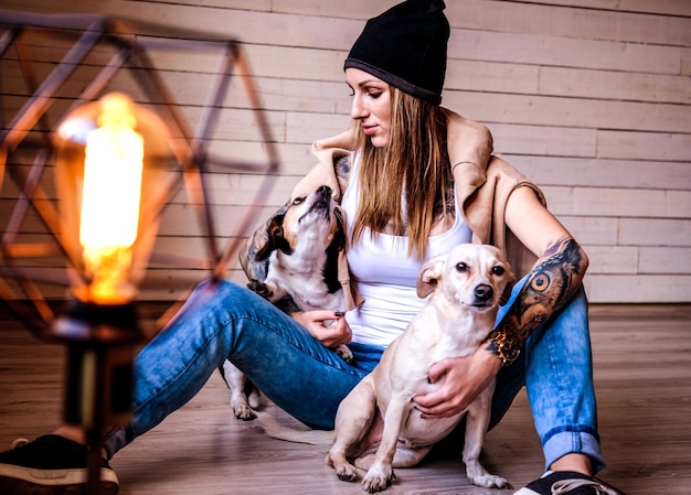 Retrato de una chica hipster tatuada jugando con sus perritos mientras se sienta en un suelo de madera.